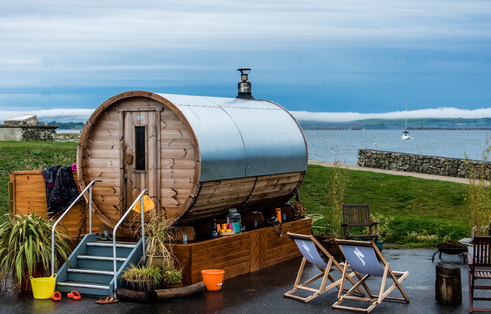 A wooden barrel sauna on the shore with deckchairs and the sea behind